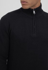 Schwarzer gestrickter Pullover mit hohem geripptem Kragen, Halb-Reißverschluss, strukturiertem Muster und Raglanärmeln.