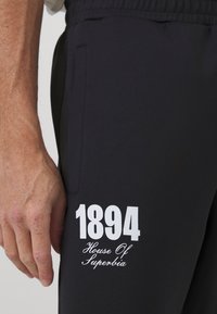 Melnas jogger bikses ar elastīgu jostas vietu. Uz kreisās puses ir balti uzraksti "1894" un "House Of Superbia", gluda auduma tekstūra.