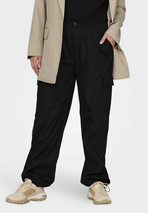 Sorte cargo bukser med elastisk talje, sidelommer og afslappet pasform, parret med beige sportssko og en lys beige blazer.