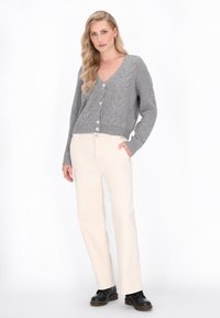 Grijze gebreide cardigan met een V-hals, voorzien van een kabelpatroon en grote knopen, gecombineerd met crèmekleurige broek en zwarte schoenen.