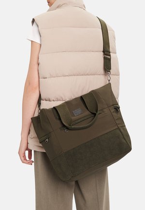 Osoba w beżowej puchowej kamizelce niesie dużą oliwkowozieloną torbę typu tote z podwójnymi uchwytami i regulowanym paskiem na ramię na plecach.