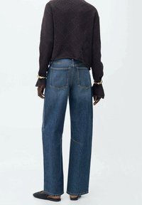 Jean large en denim foncé, avec des poches arrière et des coutures visibles. Associé à un pull noir texturé et des chaussures noires.