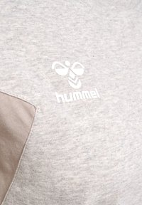 Lys grå sweatshirt med en beige detalje på skulderen og et hvidt "hummel" logo med en bi og chevron-mønster. Blød tekstur.
