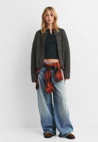 Cardigan grigio a maglia sopra un top nero corto, abbinato a jeans larghi azzurri chiari e una camicia a quadri rossa annodata in vita. Calzature marroni.