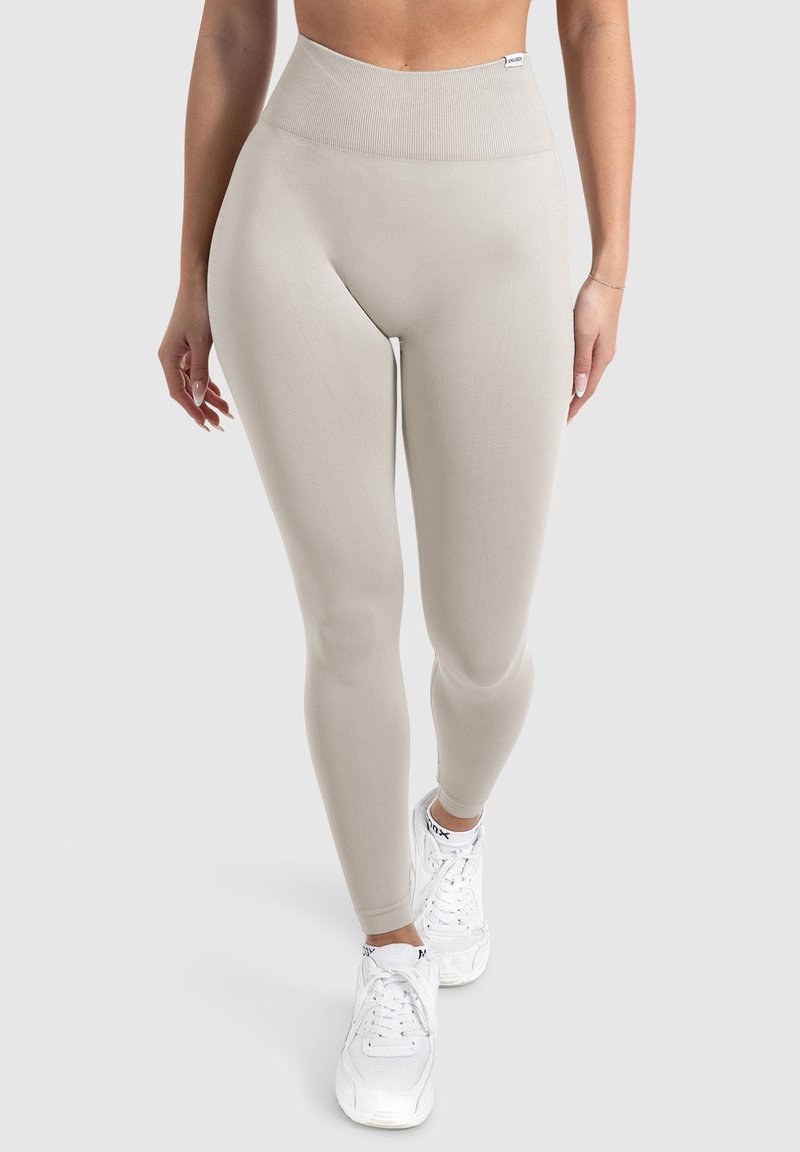 Beige high-waisted leggings van rekbaar materiaal, met een gladde textuur en een strakke pasvorm, gecombineerd met witte sportschoenen.