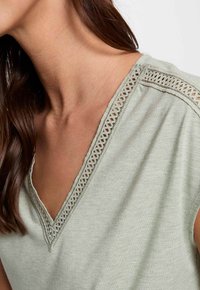 Jasnozielona bluzka w serek wykonana z miękkiego materiału, z koronkowym wykończeniem wzdłuż dekoltu i subtelnymi, połyskującymi detalami w tkaninie.