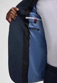 Dopasowana marynarka z teksturowanym ciemnoniebieskim zewnętrzem, kontrastową czarną podszewką oraz jasnoniebieskim wnętrzem z metką z napisem "Najlepsza włoska tkanina".