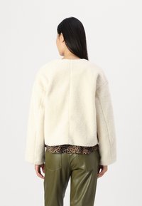 Kremfarget, teksturert fleecejakke med brede ermer og cropped design, lagdelt over en leopardmønstret topp og olivengrønne bukser.