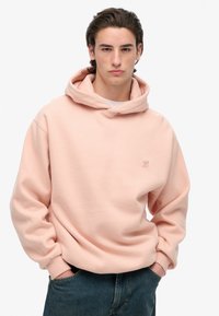 Sudadera rosa hecha de una tela suave, con un gran bolsillo, puños acanalados y un pequeño logo bordado en el pecho. Combinada con jeans de mezclilla azules.