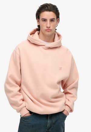 Pink hoodie made of soft fabric, featuring a large pocket, ribbed cuffs, and a small embroidered logo on the chest. Paired with blue denim.