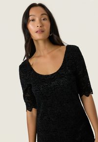 Mustat pitsimekko, jossa on kaarroke-kaula-aukko ja lyhyet, aallotetut hihat. Koristeltu kukkakuoseilla ja hienovaraisella tekstuurilla koko ajan.