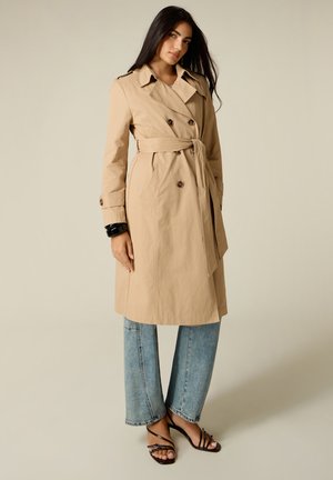 Mujer con un abrigo trench beige con cinturón sobre vaqueros azules y sandalias negras de tacón con punta abierta, de pie frente a un fondo liso.