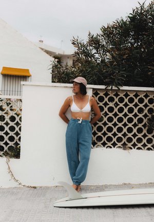 Woman in white top, blue sweatpants, and pink cap leaning against white wall, barefoot next to surfboard on tiled pavement.