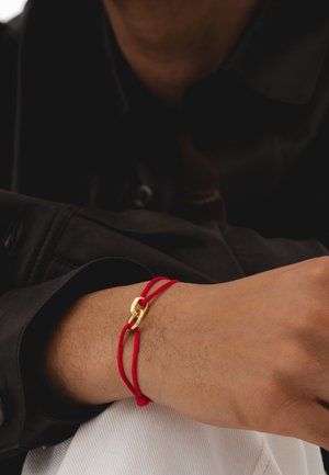 Le bracelet élastique rouge présente un fermoir en métal doré, porté au poignet. Le design est minimaliste avec des textures lisses et des couleurs contrastées.