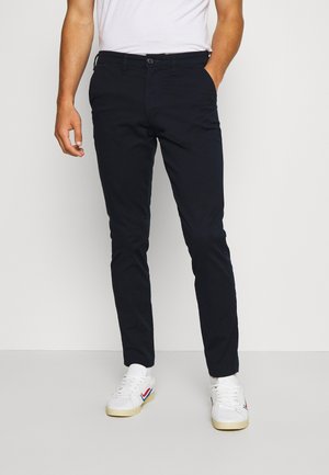 Navyblauwe chino's met een standaard pasvorm, voorzien van een sluiting met knoop aan de voorkant, riemlussen en zijzakken. Draagt men met witte sneakers.