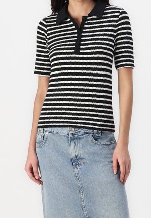 Kvinde iført en sort og hvid stribet kortærmet polo skjorte, stoppet ned i en lys blå denimnederdel med lommer og knapfront.