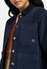 Marinblå kordjacka med knappstängning, två framfickor och logotypbroderi. Designen inkluderar en spridd krage och strukturerad tyg.