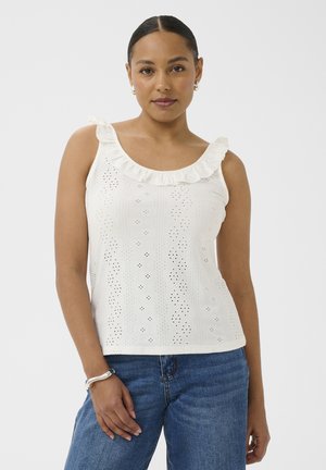 Žena oblečená v bielom bezrukávovom top s volánikmi a očkovými vzormi, v modrých džínsoch, pózujúca s jednou rukou položenou pri bokoch.