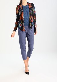Cardigan floral multicolor sobre una blusa azul, combinado con pantalones grises recortados. La modelo lleva tacones negros. Textura suave, corte relajado.
