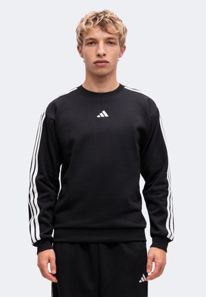 Jeune homme aux cheveux blonds courts portant un sweat Adidas noir avec des bandes blanches sur les manches et un pantalon noir assorti.