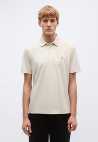 Béžové polotričko z bavlny s klasickým golierom, tromi gombíkmi, textúrovanou látkou a malou logovou nášivkou na ľavej strane hrudníka.