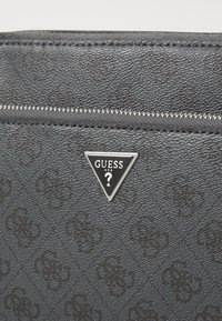 Bolso de mano negro texturizado con cierre de cremallera, patrón embossado y un logotipo triangular plateado con la marca "GUESS".