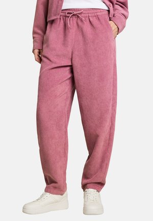 Persona che indossa pantaloni in velluto a coste rosa a vestibilità ampia con vita elastica e sneakers bianche, in piedi con una mano in tasca.