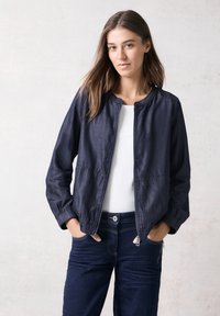 Marineblau leichte Bomberjacke mit rundem Ausschnitt, Reißverschluss vorne und elastischen Bündchen, kombiniert mit einem weißen Oberteil und dunklen Jeans. Glatte Textur.