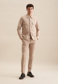 Beige skjorte med krave og to frontlommer, parret med matchende tapered bukser og sorte snørebåndssko. Glat tekstur, skræddersyet pasform.