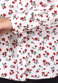 Witte bloemenshirt met een patroon van rode bloemen, groene bladeren en knoopsluiting aan de voorkant. De stof lijkt lichtgewicht met een gladde textuur.