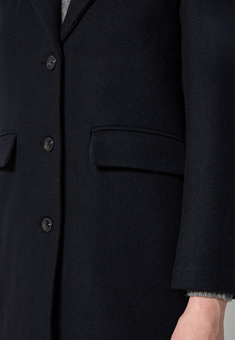 Manteau en laine noire au design structuré, avec deux poches avant, trois boutons et un poignet gris texturé visible en dessous.