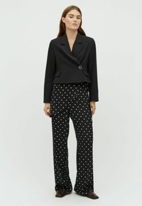 Čierny skrátený blazer s jedným strieborným gombíkom a dvoma prednými vreckami, skombinovaný s vysokými čiernymi nohavicami s bielymi bodkami.