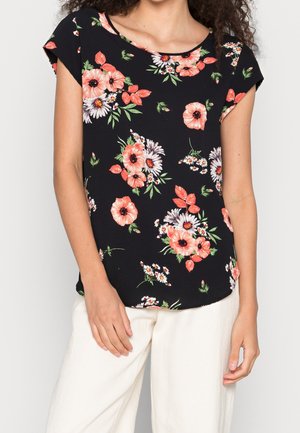 Vrouw die een zwart kortemouwtop met oranje-witte bloemenprint en een witte broek draagt, staat met ontspannen armen langs haar zij.