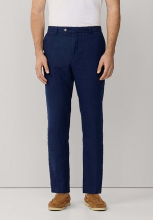 Homme portant un pantalon bleu marine sur mesure, une chemise blanche et des mocassins en daim beige, debout devant un fond uni gris clair.