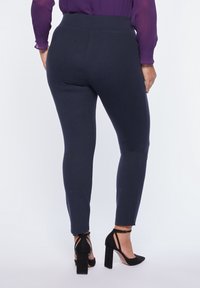 Marinblå leggings med en mjuk textur och elastisk midja, i kombination med svarta klackskor, visade bakifrån.