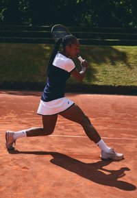 Tennis spiller i en marineblå og hvid kjole, der svinger en ketcher på grusbanen. Iført grå sko og hvide strømper, med flettet hår i bevægelse.