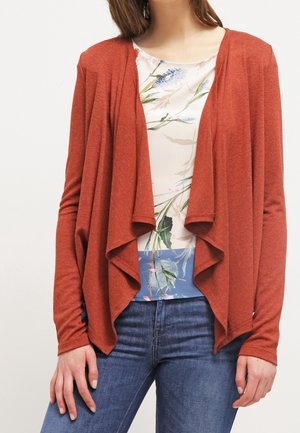 Kvinde iført en rustfarvet åben cardigan over en blomsterbluse og blå jeans, stående mod en ensfarvet baggrund.