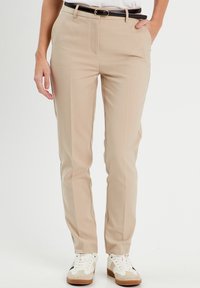 Pantalon beige sur mesure avec une texture lisse, doté d'une fermeture éclair frontale et de deux poches latérales, porté avec une ceinture noire et des baskets blanches.