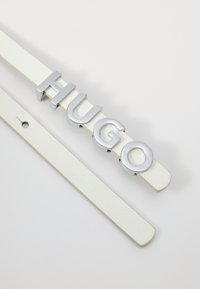 Weißer Leder Gürtel mit einem silbernen, metallischen "HUGO" Logo, flaches Design, strukturierte Oberfläche, mit einem Loch zur verstellbaren Befestigung.
