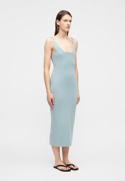 Γυναίκα με αμάνικο μίντι φόρεμα ανοιχτού μπλε χρώματος με διακοσμητικά κουμπιά κατά μήκος του λαιμού, στέκεται με μαύρα σαγιονάρες σε λευκό φόντο.