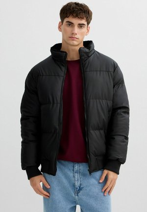 Chaqueta acolchada negra con cuello alto, cierre de cremallera, puños acanalados y textura suave. Se lleva sobre una camisa burdeos y unos vaqueros azul claro.