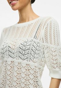 Maglione bianco lavorato a maglia con intricati motivi a traforato, punti strutturati e scollatura rotonda. Maniche corte con polsini a coste.