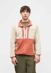 Νέος άνδρας που φοράει ένα μπεζ και κοραλλί φλις τζάκετ Columbia με φερμουάρ πάνω από ένα καφέ πουκάμισο και χακί παντελόνια cargo, στέκεται σε άσπρο φόντο.