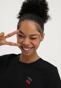 Mlada žena s kovrčavom kosom u punđi smiješi se, namiguje i drži znak mira blizu oka, nosi crnu majicu s logotipom trešnje.