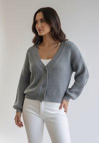 Szary sweter na drutach z dekoltem w serek, długimi rękawami i trzema guzikami z przodu. Nosi się go na białą bluzkę i łączy z białymi spodniami.