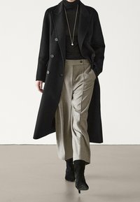 Manteau en laine noir croisé porté sur un col roulé noir, associé à un pantalon beige plissé et des bottines noires, sur un fond neutre.
