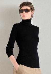 Černý žebrovaný rolák s dlouhými rukávy, ve fitted siluetě. Stylizováno s oversized hnědými slunečními brýlemi a světlým pozadím.