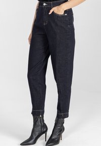 Ciemnoniebieskie spodnie dżinsowe z wysokim stanem, zwężającymi się nogawkami i przednimi kieszeniami. Posiadają kontrastowe przeszycia i czarne, spiczaste botki na kostkę.