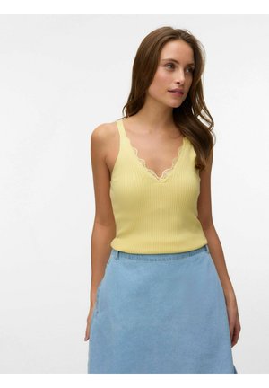 Ženska z dolgimi rjavimi lasmi, oblečena v rumen rebrast top z čipkastim robom in svetlomodro krilo z visokim pasom, gleda proti svoji desni.