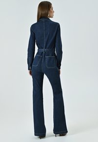 Dunkelblauer Denim-Overall mit strukturiertem Kragen, langen Ärmeln, tailliertem Bund und ausgestellten Beinen. Die Rückseite verfügt über zwei Taschen.